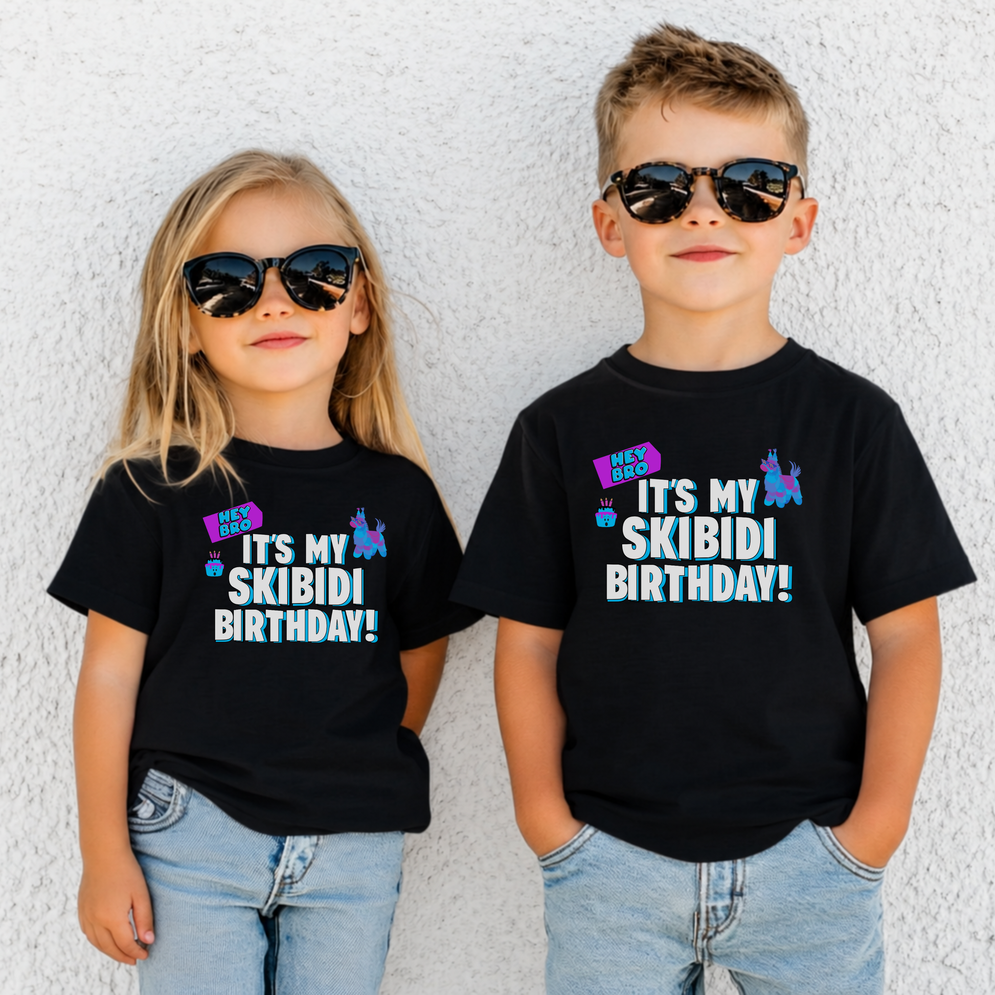 A young girl and boy wearing matching black graphic t-shirts that read 'IT'S MY SKIBIDI BIRTHDAY!' in white, blue, and pink, styled with light-wash jeans and black sunglasses against a white textured wall. Skibidi birthday party shirt for kids featuring bold white and blue birthday lettering, colorful playful graphics, and a funny Gen Alpha theme on a black Bella Canvas youth t shirt