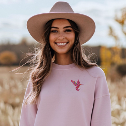 Light pink Gildan crewneck sweatshirt featuring a colorful embroidered hummingbird on the chest. The soft pink background creates a gentle contrast with the vibrant bird embroidery and gives the classic sweatshirt a feminine, nature inspired feel. This casual crewneck is easy to style with jeans or leggings and makes a thoughtful gift for a hummingbird lover, bird lover, gardener, nature enthusiast, mom, or friend.