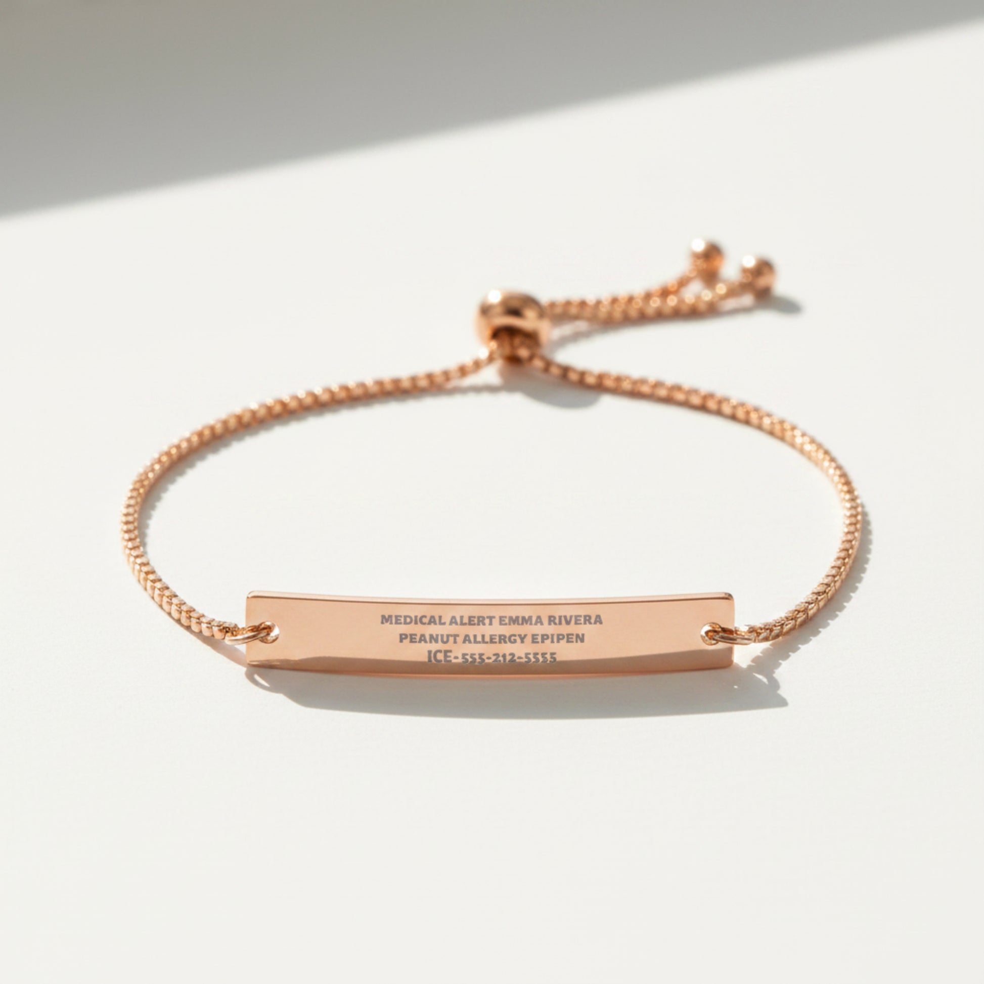 A rose gold medical alert bracelet featuring a horizontal bar engraved with personal and allergy information, suspended on a matching adjustable chain with a sliding clasp, shown on a plain white background.