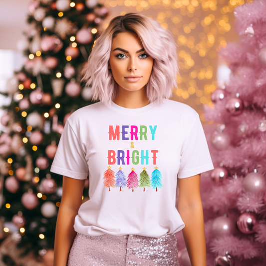 Woman wearing a Merry and Bright Christmas shirt featuring colorful rainbow trees and bold festive lettering on a soft Bella Canvas 3001 tee, styled in a cozy holiday setting with pink Christmas trees and warm lights, perfect for Christmas outfit ideas, family photos, teacher gifts, cute holiday shirts for women, unisex Christmas shirt, festive graphic tee, colorful holiday apparel, boutique style Christmas clothing