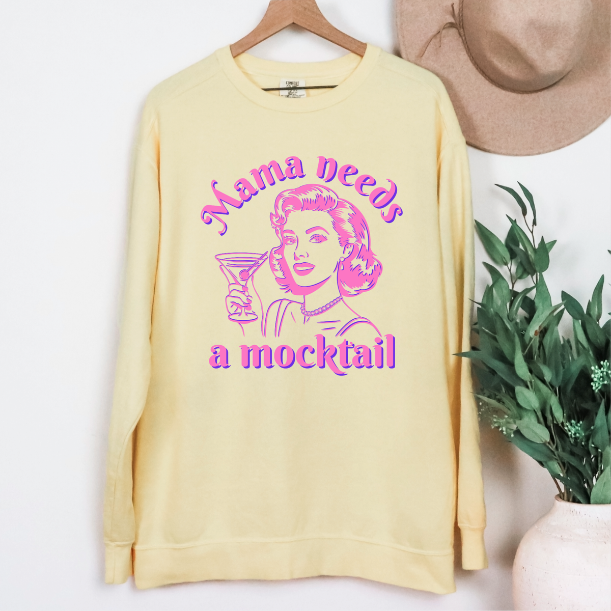 Retro pop art Mama Needs a Mocktail sweatshirt shown as a playful gift idea for a mom who loves mocktails and colorful vintage inspired designs. The graphic features an illustrated woman holding a cocktail glass surrounded by bold pink and blue lettering. This Comfort Colors crewneck makes a fun gift for expecting moms, sober curious friends, mocktail lovers, Mother's Day, birthdays, baby showers, brunch gatherings, or simply surprising a funny and fabulous mama.