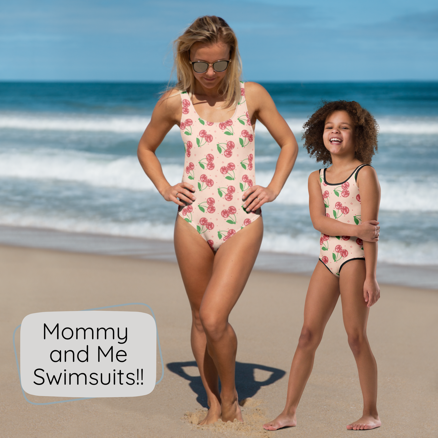 A woman and a child wearing matching disco cherry print one piece swimsuits stand on a sandy beach with the ocean in the background. A text bubble in the lower left reads, 'Mommy and Me Swimsuits!!'.
