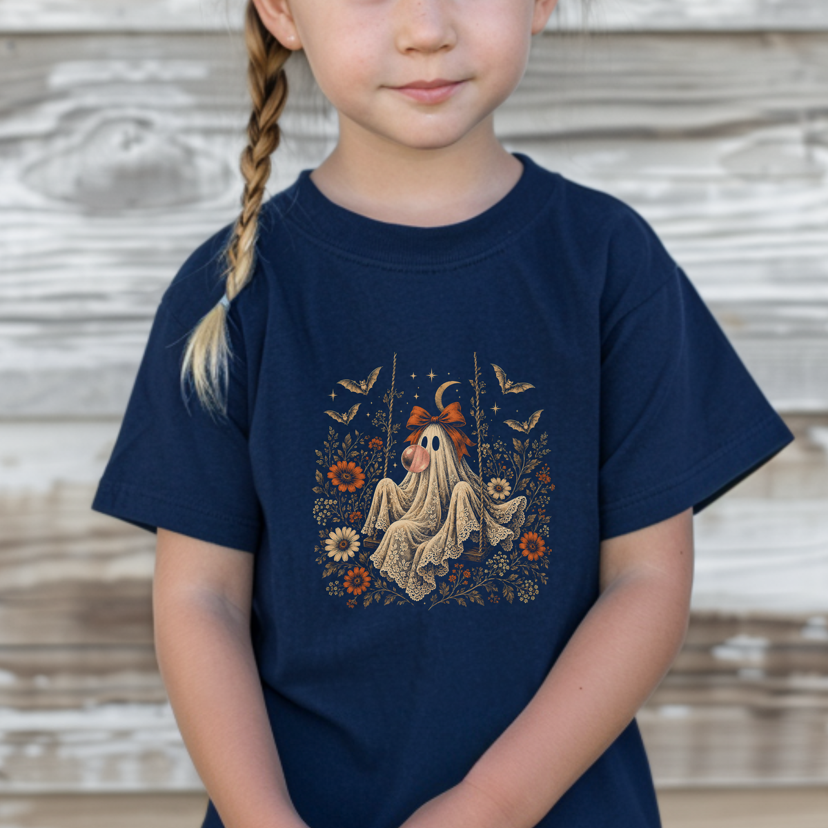 Toddler wearing a navy Halloween T shirt featuring a vintage inspired ghost blowing a bubble with bubble gum, surrounded by orange flowers, botanical foliage, bats, stars, lace and a bow. A cute toddler Halloween shirt for pumpkin patches, fall outings, Halloween parties and spooky season fun.