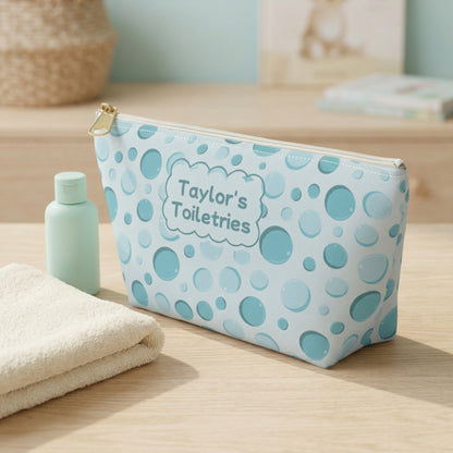 Side angle view of a personalized aqua bubble print kids toiletry pouch showing its compact depth, colorful bubble pattern, white zipper, and gold zipper pull. Styled on a light wood surface with an ivory hand towel and a small travel toiletry bottle, this custom zippered bag is ideal for kids, tweens, and teens packing toiletries for sleepovers, summer camp, and family vacations.