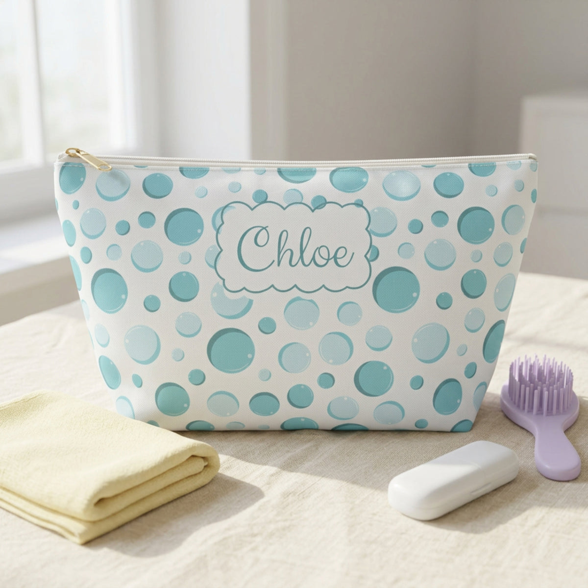 Personalized aqua bubble print kids toiletry pouch featuring a colorful bubble pattern, scalloped name label reading Chloe, white zipper, and gold zipper pull. Styled on an ivory linen surface with a pastel washcloth, lavender hairbrush, and travel toothbrush case, this compact personalized bag is ideal for kids, tweens, and teens heading to sleepovers, summer camp, and family vacations.