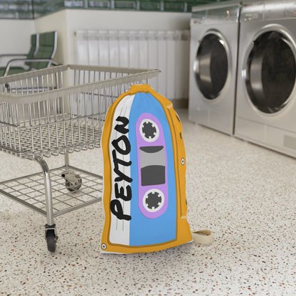 Personalized orange laundry bag featuring a colorful cassette tape pattern and custom name printed across the fabric. The bag stands upright with a drawstring closure at the top and a woven shoulder strap for easy carrying. Retro music inspired artwork covers the surface in shades of orange, yellow, blue, and teal. Spacious 18 x 29 inch design is ideal for storing clothing, towels, bedding, or camp essentials while adding a playful vintage touch to everyday organization.