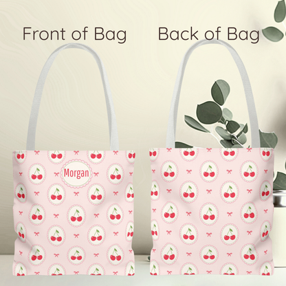 A pink tote bag shown from both front and back, with a repeating cherry print and a circular patch reading “Morgan” on the front. The image includes text labels “Front of Bag” and “Back of Bag,” with a soft beige background and decorative leaves on the right. Personalized pink tote bag decorated with repeating cherry medallions and delicate bows on a soft blush background. This Kawaii inspired cherry aesthetic accessory has a playful Cutecore and Coquette feel and makes a cute teacher gift, tween girl gift.