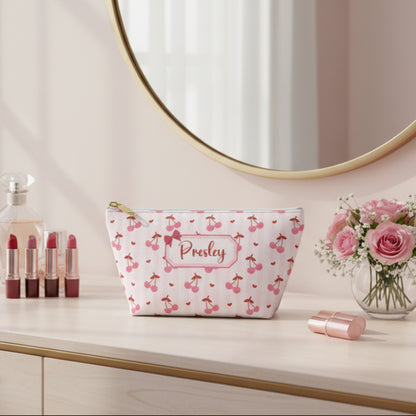 Custom cherry cosmetic pouch featuring an all over cherry print on a pastel pink background with a personalized name label in the center. This coquette inspired makeup bag includes a structured T bottom design, white zipper closure, and gold zipper pull for a polished feminine look. Perfect for storing makeup brushes, skincare, hair accessories, travel items, school supplies, and purse essentials. Cute personalized gift idea for girls, teens, moms, bridesmaids, and cherry lovers.