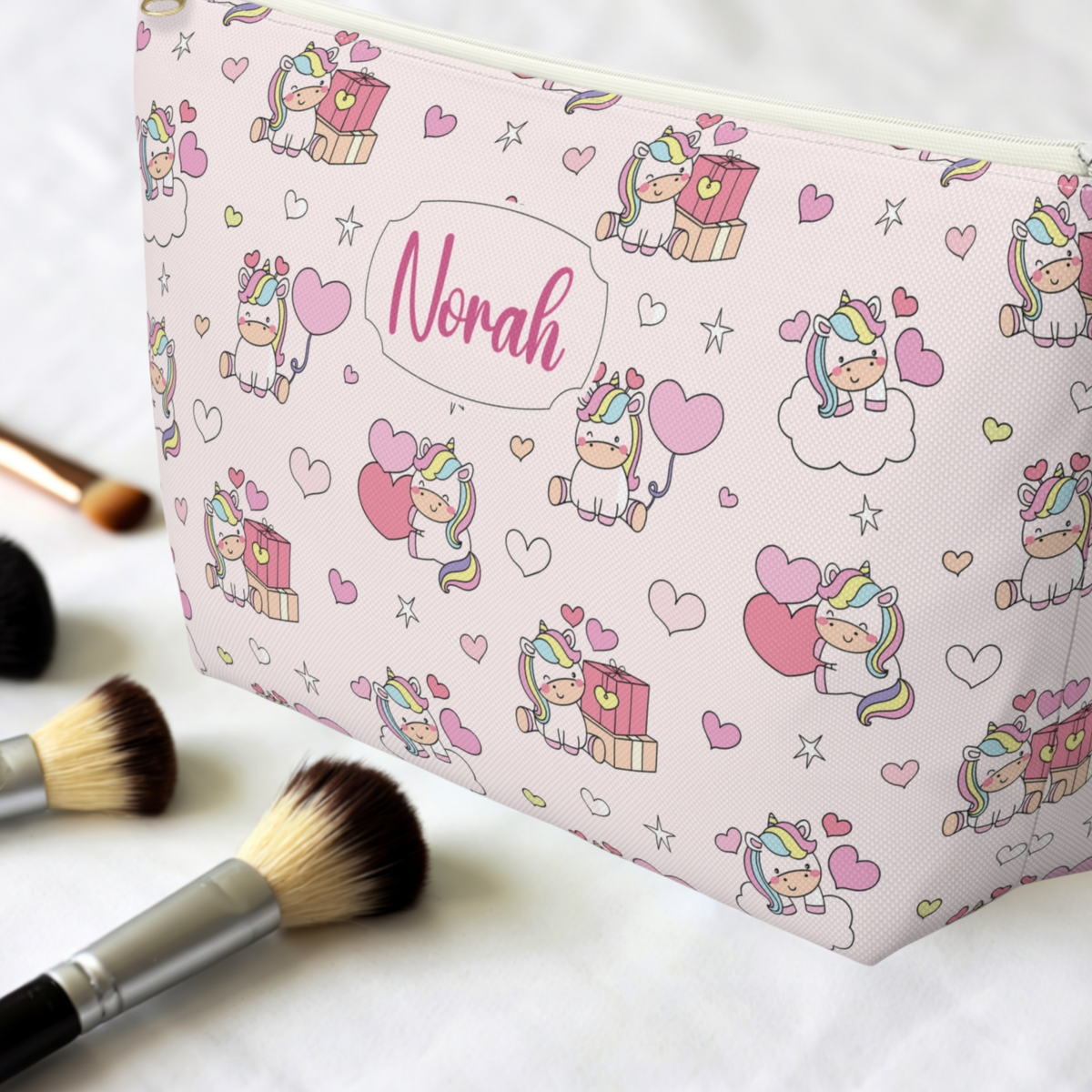 A close up view of the personalized unicorn zipper pouch next to makeup brushes shows the colorful pastel rainbow print and custom name detail. The whimsical unicorn design brings a playful cutecore and pastel aesthetic to a practical girls makeup bag. Its compact shape makes it useful for cosmetics, brushes, beauty accessories and other small essentials at home or while traveling.