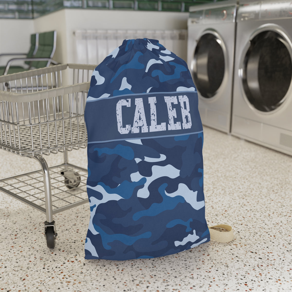 A blue camouflage drawstring laundry bag with the name 'CALEB' printed in white across the center, standing on a speckled floor next to a shopping cart in a laundry room with washing machines in the background.