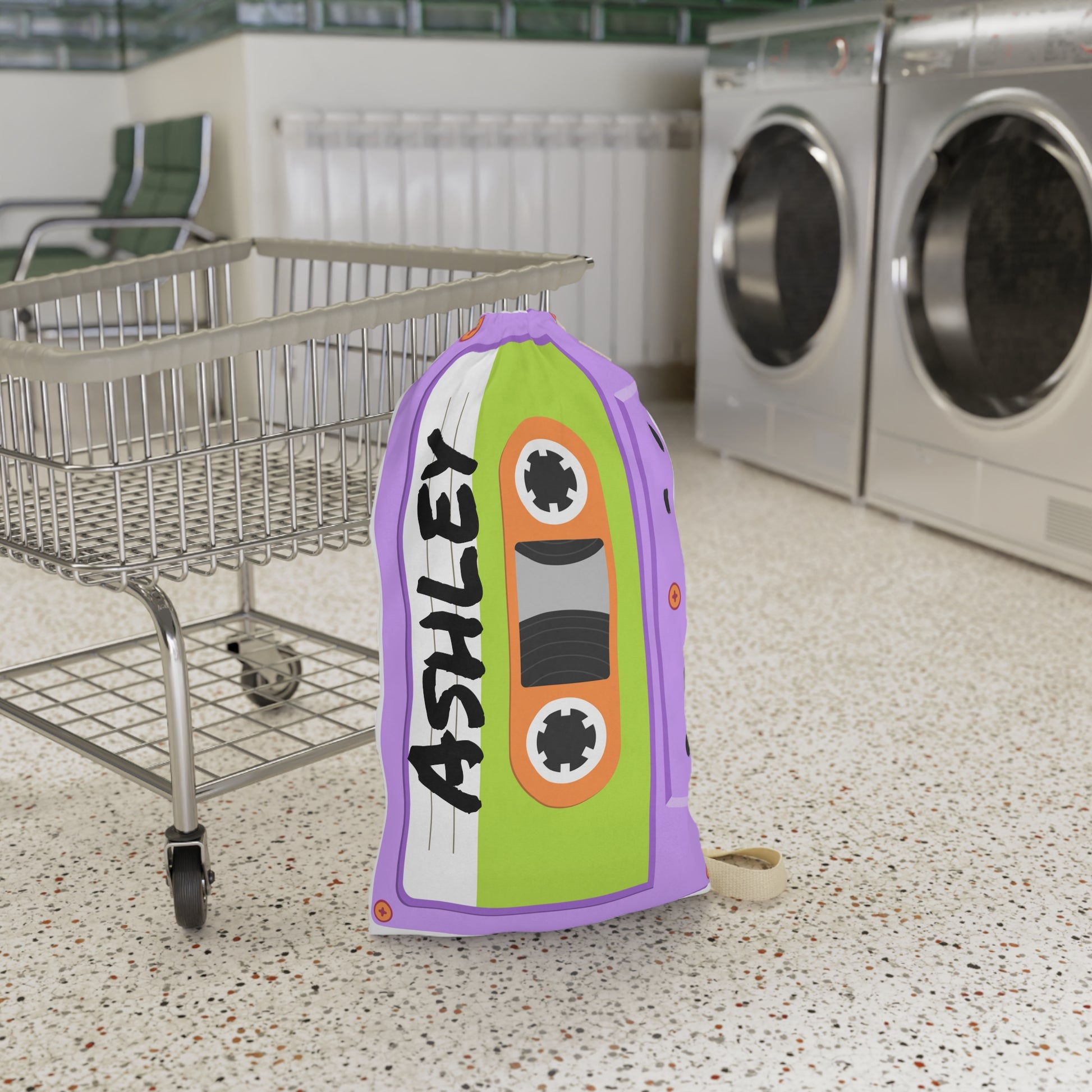 Personalized purple laundry bag featuring a colorful cassette tape pattern and custom name printed across the fabric. The bag stands upright with a drawstring closure at the top and a woven shoulder strap for easy carrying. Retro music inspired artwork covers the entire surface in shades of purple, pink, blue, and teal. Spacious design is ideal for storing clothing, towels, bedding, or camp essentials while adding a playful vintage touch to everyday organization.