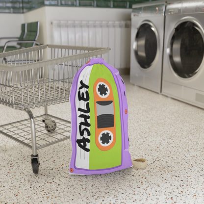 Personalized purple laundry bag featuring a colorful cassette tape pattern and custom name printed across the fabric. The bag stands upright with a drawstring closure at the top and a woven shoulder strap for easy carrying. Retro music inspired artwork covers the entire surface in shades of purple, pink, blue, and teal. Spacious design is ideal for storing clothing, towels, bedding, or camp essentials while adding a playful vintage touch to everyday organization.