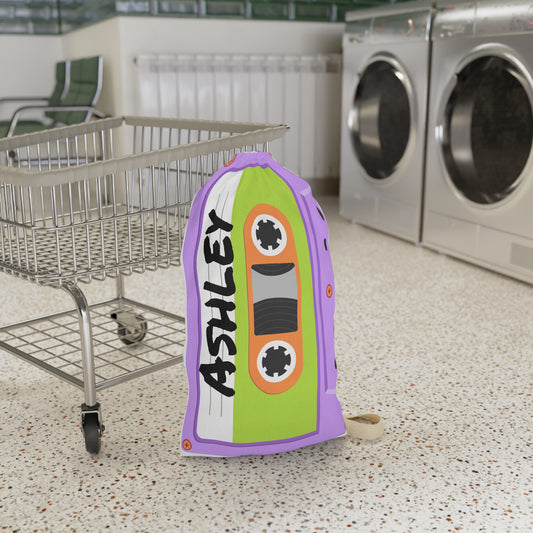 Personalized purple laundry bag featuring a colorful cassette tape pattern and custom name printed across the fabric. The bag stands upright with a drawstring closure at the top and a woven shoulder strap for easy carrying. Retro music inspired artwork covers the entire surface in shades of purple, pink, blue, and teal. Spacious design is ideal for storing clothing, towels, bedding, or camp essentials while adding a playful vintage touch to everyday organization.