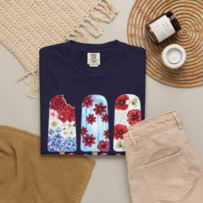 A navy blue folded T-shirt with a graphic of three popsicle shaped floral designs in red, blue, and white, styled in a flat lay with beige pants, a woven blanket, a candle jar, and a small cup on a neutral background. Personalized women's floral patriotic shirt printed on a Comfort Colors 1717 tee, featuring three red white and blue floral popsicles for summer celebrations
