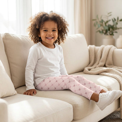 Toddler girl sitting comfortably on a cream sofa wearing pink heart leggings with a white long sleeve shirt. The stretchy full length leggings feature a high rise elastic waistband, soft pink background, and delicate diamond lattice pattern with scattered hearts. Available in sizes 2T through 7, these girls leggings are a cute choice for relaxing at home, playdates, preschool outfits, and everyday casual style.
