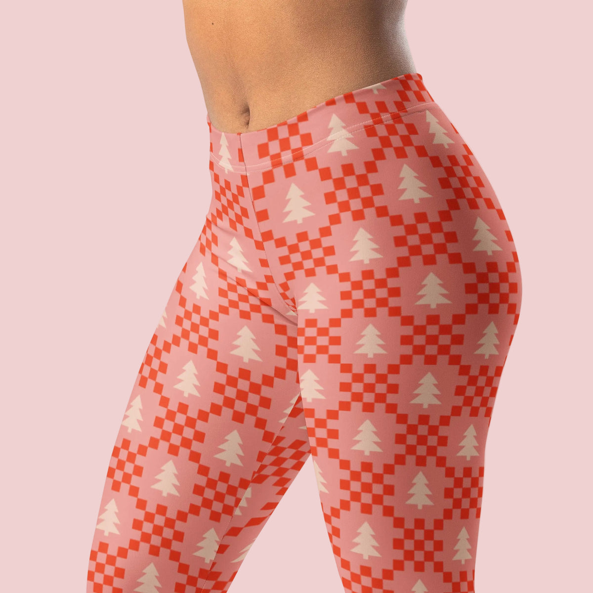 A close view of pink, red, and cream Christmas leggings showing the repeating tree and geometric pattern across the fabric. The soft pink background gives this holiday design a feminine and playful look, while the red details add a festive pop of color. These women's leggings are a fun alternative to traditional red and green Christmas clothing and can be styled throughout the holiday season.