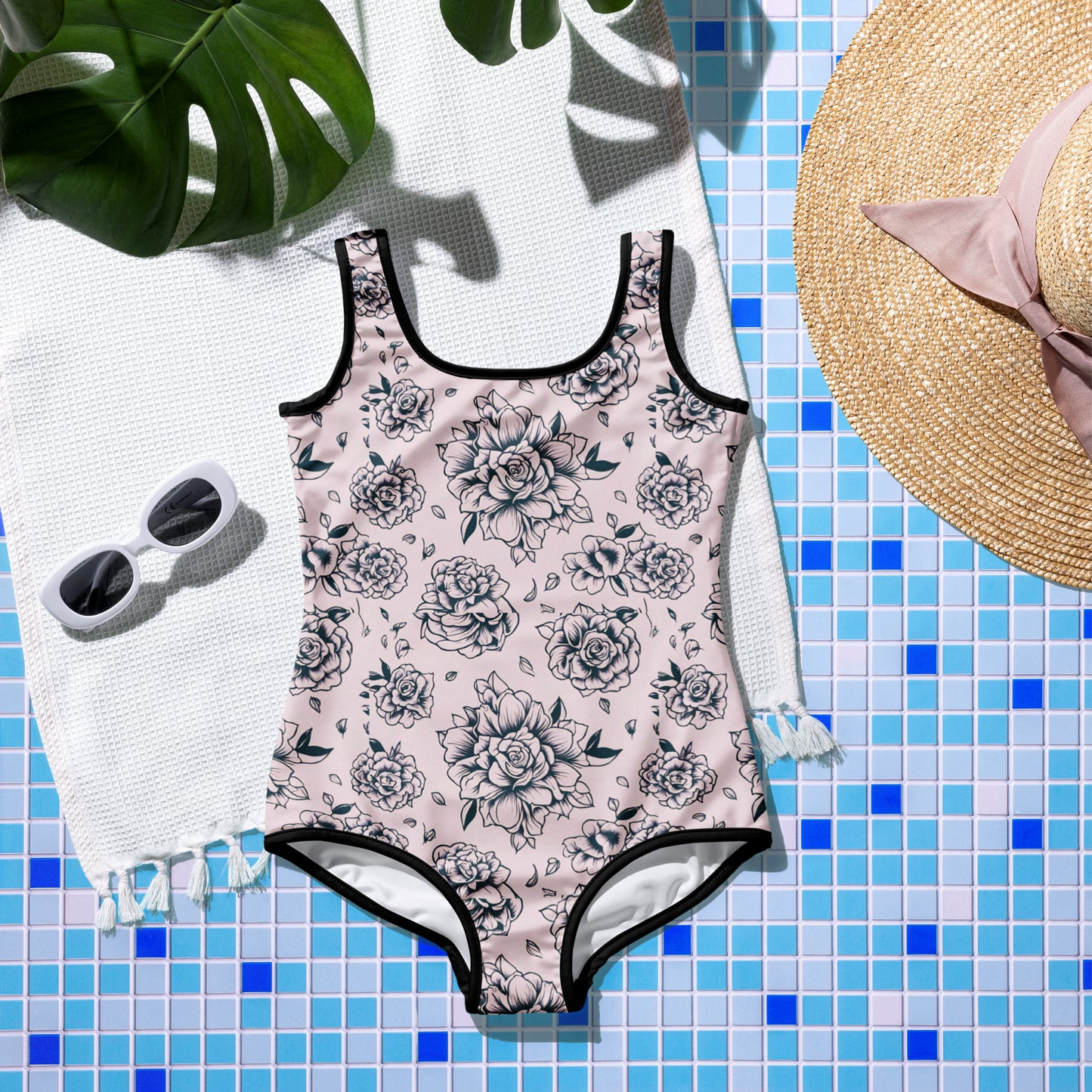 A pink one-piece swimsuit with a black tattoo roses pattern, displayed on a flat surface with a straw hat, sunglasses, and a towel in the background, set against a poolside or beach-like setting.