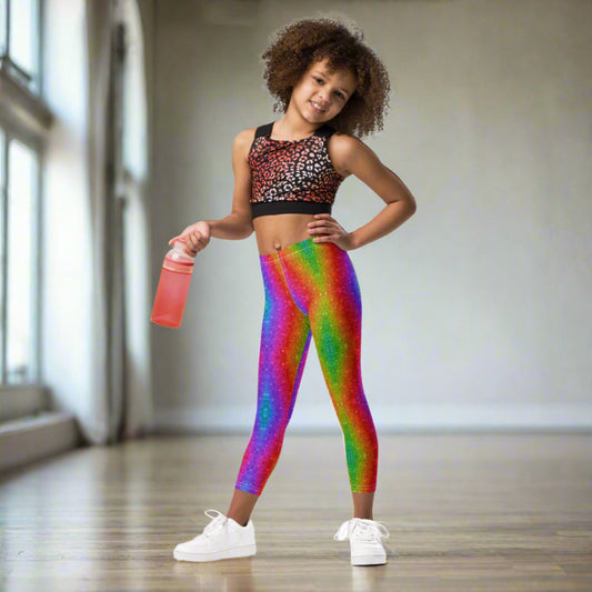 Young girl wearing rainbow glitter holographic swirl leggings in a vibrant multicolor pattern, showing the full length fit and colorful faux glitter effect. These stretchy girls leggings are available in toddler and tween sizes 2T–20 and are designed for comfortable movement during dance, play, school, parties, festivals, parades, and everyday wear. The bright rainbow design also makes them fun for colorful family matching outfits.