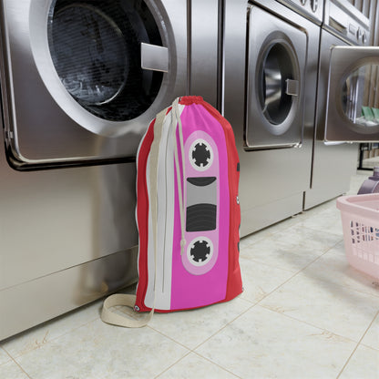 Custom laundry sack decorated with a vibrant red background, bright pink cassette tape pattern, and personalized name. The roomy 18 x 29 inch design includes a secure drawstring closure and sturdy shoulder strap for carrying clothes between bedrooms, dorm rooms, camp cabins, or laundromats. Colorful retro graphics create a playful nostalgic appearance while helping keep laundry organized and easy to transport.