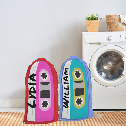 Red and blue personalized laundry bags covered in colorful cassette tape artwork. A custom name is incorporated into the print, creating a unique storage solution for everyday use. The 18 x 29 inch bag features a woven carrying strap and gathered drawstring closure. Ideal for storing clothing, linens, towels, camp gear, and travel essentials while bringing retro style to bedrooms and dorm rooms.