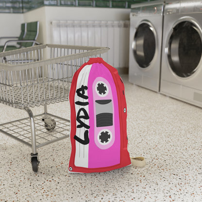Personalized red laundry bag featuring colorful cassette tape illustrations accented with bright pink details and a custom name printed across the fabric. The 18 x 29 inch bag includes a drawstring closure at the top and a woven shoulder strap for convenient carrying. Retro music inspired artwork covers the surface, creating a fun vintage look. Spacious design provides room for clothing, towels, bedding, and camp essentials while adding personality to everyday organization.