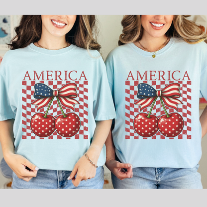 Two women weating the retro Americana Comfort Colors shirt featuring playful cherry coquette design. America text crowns two bright cherries decorated in white stars and connected by a large flag inspired bow. Distressed checkerboard backdrop gives classic summer race track vibes with a boutique look. Relaxed cotton tee with soft garment dyed texture for all day comfort. Perfect for Fourth of July parties, beach boardwalk nights, county fairs, and patriotic photo shoots.
