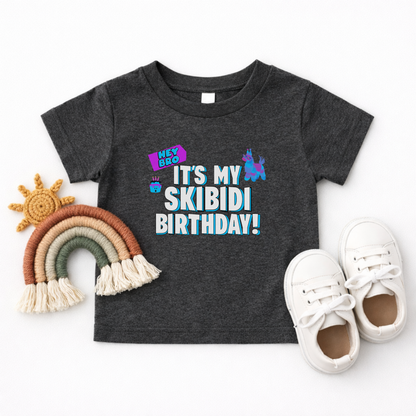 A toddler's dark heather gray t-shirt with a colorful 'IT'S MY SKIBIDI BIRTHDAY!' graphic, styled in a flat lay with white sneakers, a woven rainbow, and a woven sun on a white background. Funny toddler graphic shirt for a Skibidi birthday party featuring colorful birthday artwork, bold white and blue lettering, and a playful Gen Alpha design for a fun toddler birthday celebration