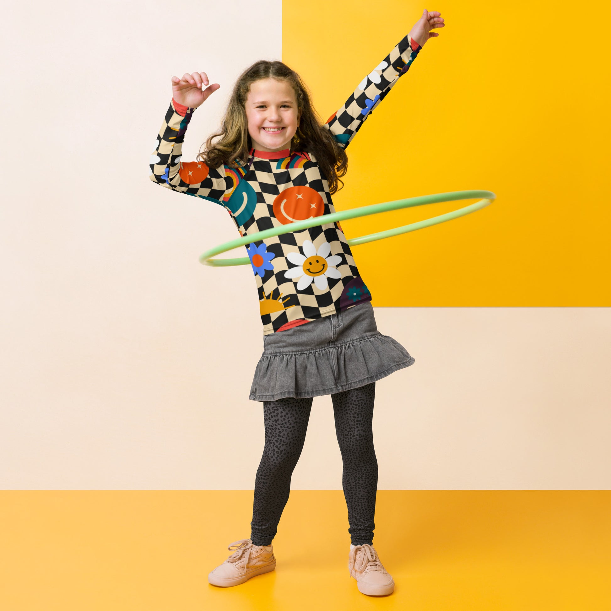 A child in a colorful checkered rash guard with a green hoop, on a yellow background, with a gray skirt and beige shoes.