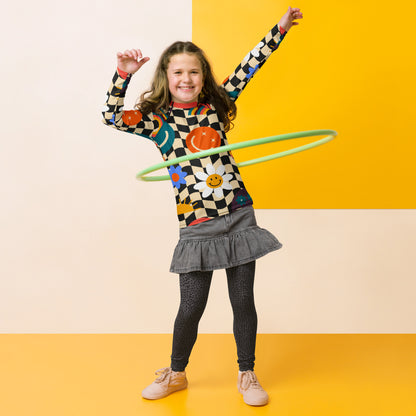A child in a colorful checkered rash guard with a green hoop, on a yellow background, with a gray skirt and beige shoes.