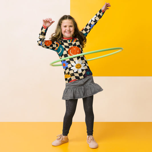 A child in a colorful checkered rash guard with a green hoop, on a yellow background, with a gray skirt and beige shoes.