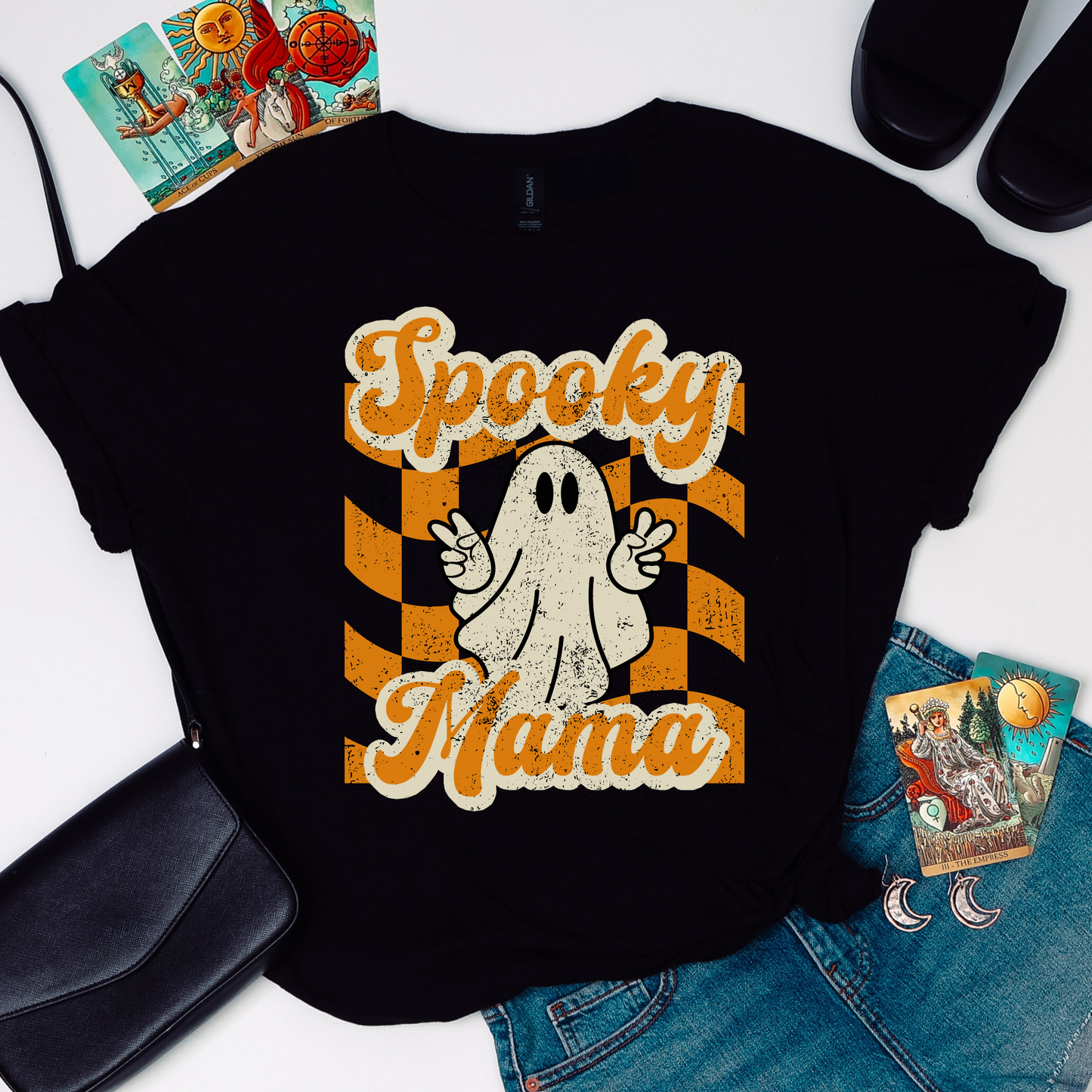 Spooky Mama Halloween tshirt in black featuring an adorable ghost giving peace signs on an orange checkerboard background for a fun retro fall look. Printed on a lightweight Gildan 64000 tee with soft all day comfort. Perfect Halloween shirt for moms during October parties, school pickup, candy runs, pumpkin patch days, and family photos. Great gift for moms who love cute spooky fashion. Matching Spooky Mini child tee available. Free U.S. Shipping.