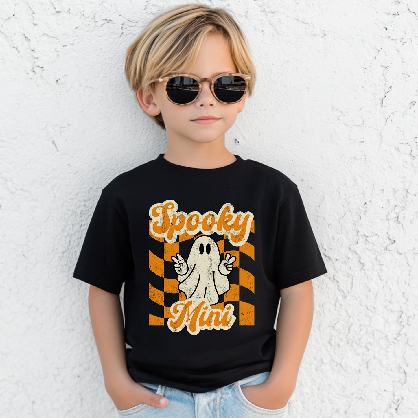 Youth Halloween shirt featuring Spooky Mini ghost design with cute peace sign hand gesture over orange checkered retro background printed on a black Bella Canvas 3001Y cotton tee. Perfect for youth Halloween outfits, school spirit days, pumpkin patch visits, trick or treating, and fall family photos. Coordinates with toddler Spooky Mini tee and adult Spooky Mama shirt for matching mommy and me Halloween outfits and family Halloween clothing sets.