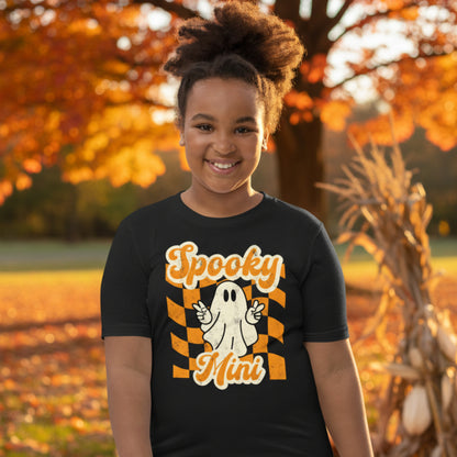 Youth Halloween t shirt featuring Spooky Mini ghost graphic with peace sign hand gesture and orange checkered background printed on Bella Canvas 3001Y black tee. Designed for youth fall outfits, Halloween school parties, pumpkin patch visits, trick or treating, and seasonal family photos. Matches toddler Spooky Mini shirt and adult Spooky Mama tee for coordinated mommy and me and full family Halloween matching outfits.