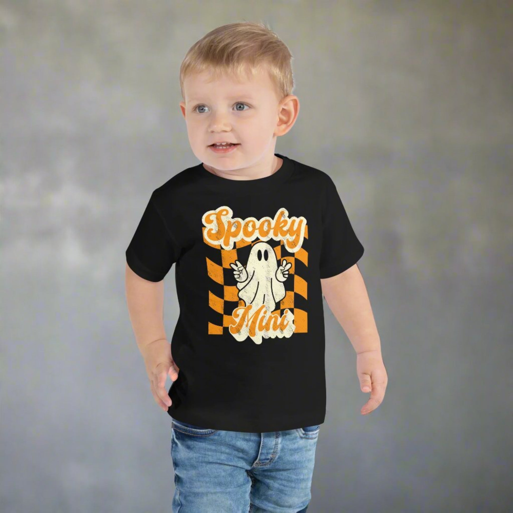 Adorable Spooky Mini toddler Halloween shirt featuring cute ghost with peace sign hand gesture on orange checkered retro style background printed on Bella Canvas 3001T cotton tee. Designed for comfort and style during fall season activities including pumpkin patch visits, Halloween parties, school events, and trick or treating. Soft fabric makes it perfect for toddlers, and matching Spooky Mama shirt completes family coordinated outfits.