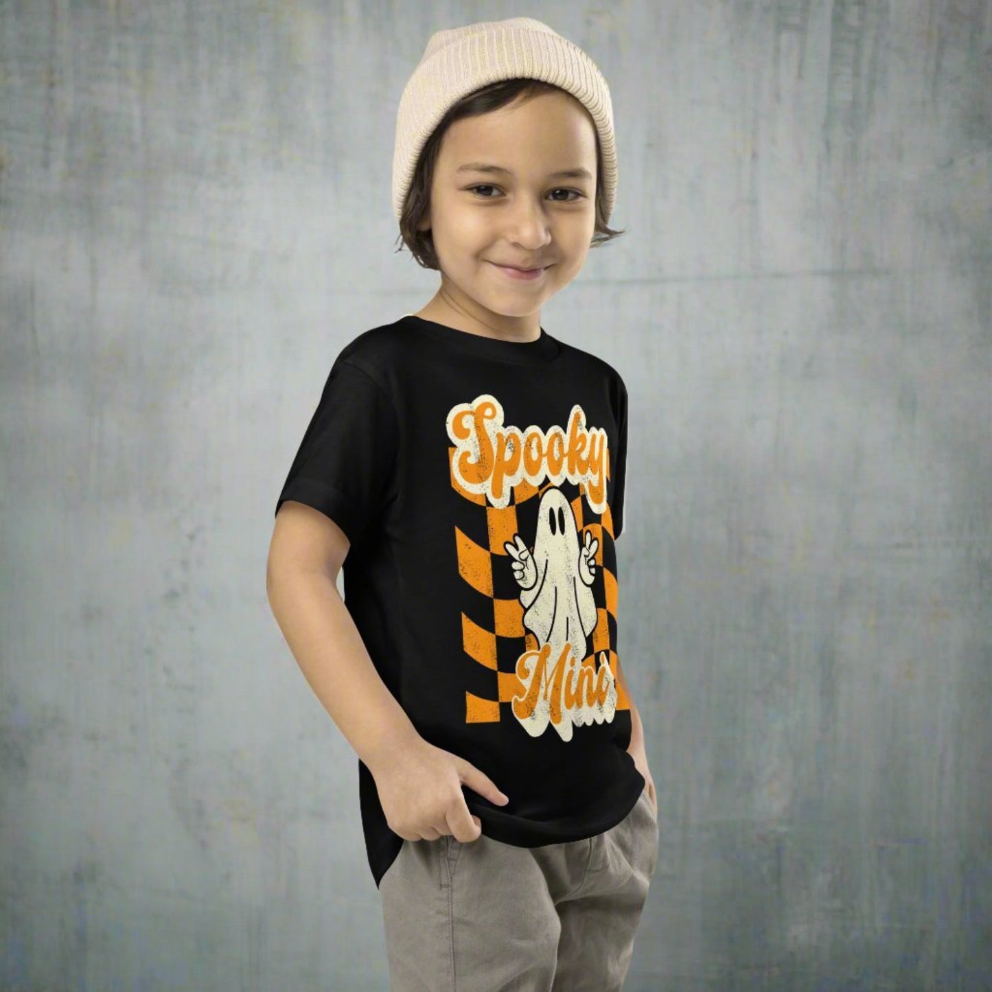 Halloween toddler shirt featuring Spooky Mini ghost graphic with peace sign over retro orange checkered background design printed on a soft Bella Canvas 3001T cotton tee. Perfect for spooky season outfits, trick or treating, pumpkin patch photos, school celebrations, and fall family events. Lightweight and breathable for comfort all day long. Matching Mama and Mini tees available featuring coordinating Spooky Mama design for family matching looks.