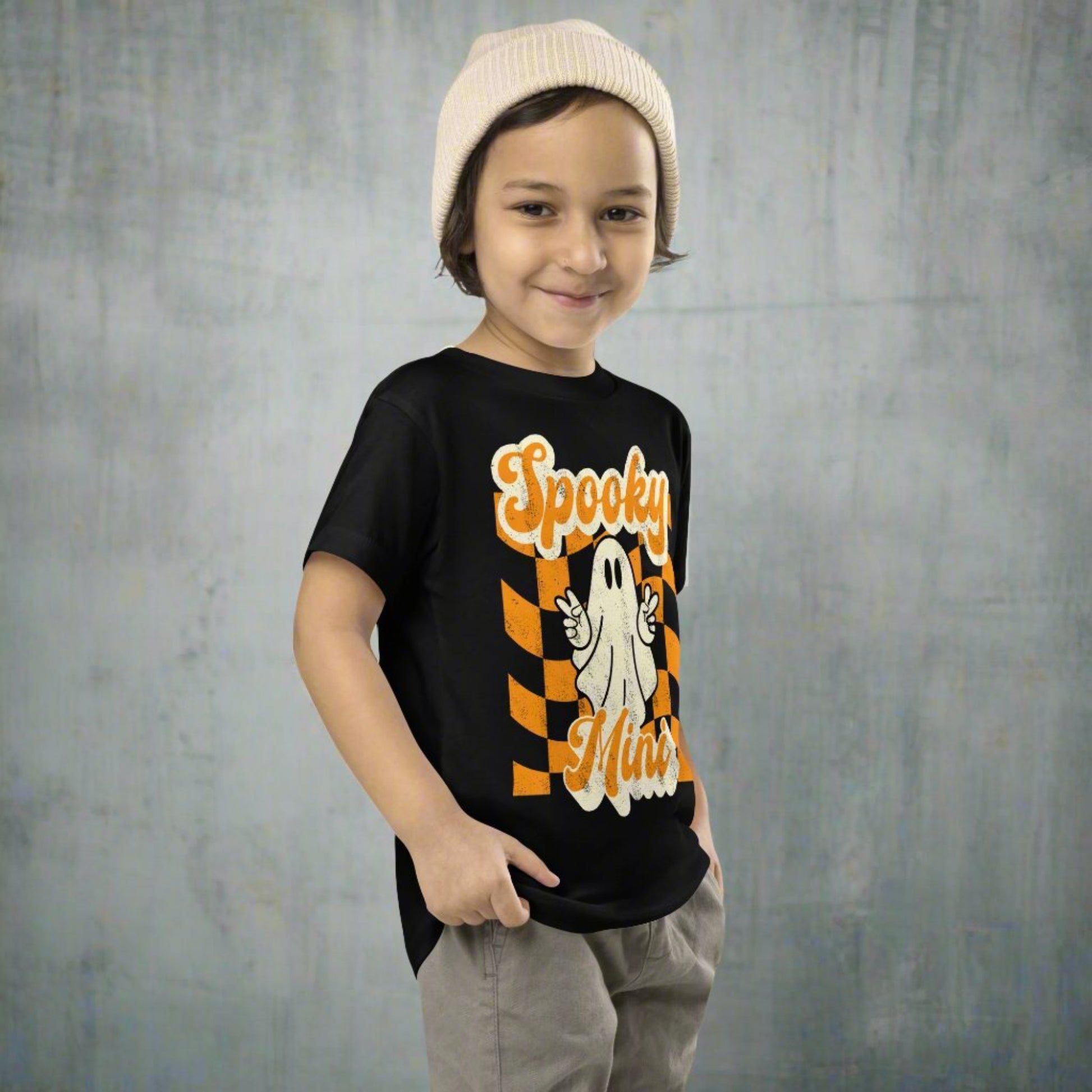 Halloween toddler shirt featuring Spooky Mini ghost graphic with peace sign over retro orange checkered background design printed on a soft Bella Canvas 3001T cotton tee. Perfect for spooky season outfits, trick or treating, pumpkin patch photos, school celebrations, and fall family events. Lightweight and breathable for comfort all day long. Matching Mama and Mini tees available featuring coordinating Spooky Mama design for family matching looks.