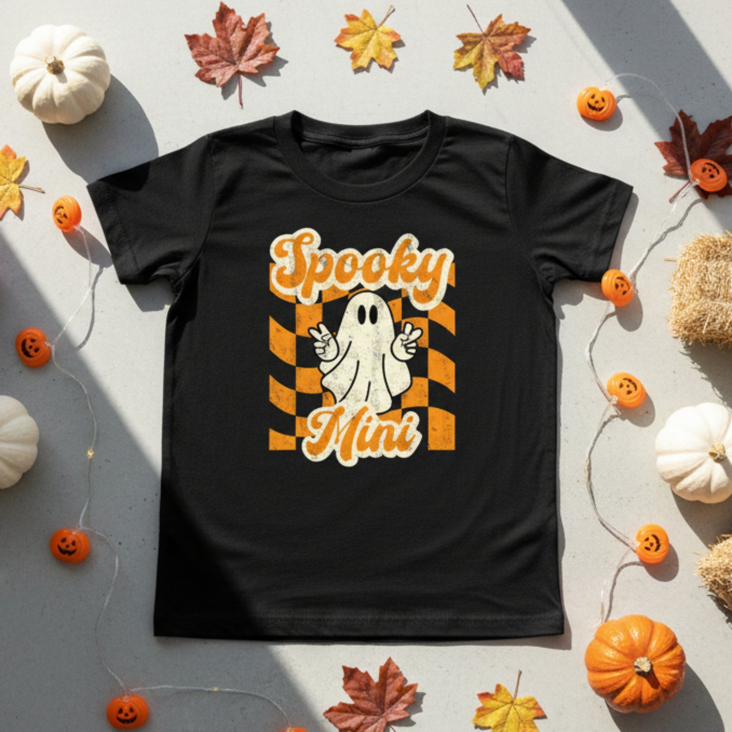 Black youth Halloween shirt featuring Spooky Mini design with cute ghost making peace sign over orange checkered retro aesthetic background printed on Bella Canvas 3001Y tee. Perfect for youth Halloween outfits, fall wardrobe styling, school spirit days, pumpkin patch photos, and trick or treating. Part of matching family collection with toddler Spooky Mini tee and Spooky Mama adult shirt for coordinated Halloween family outfits and photos.
