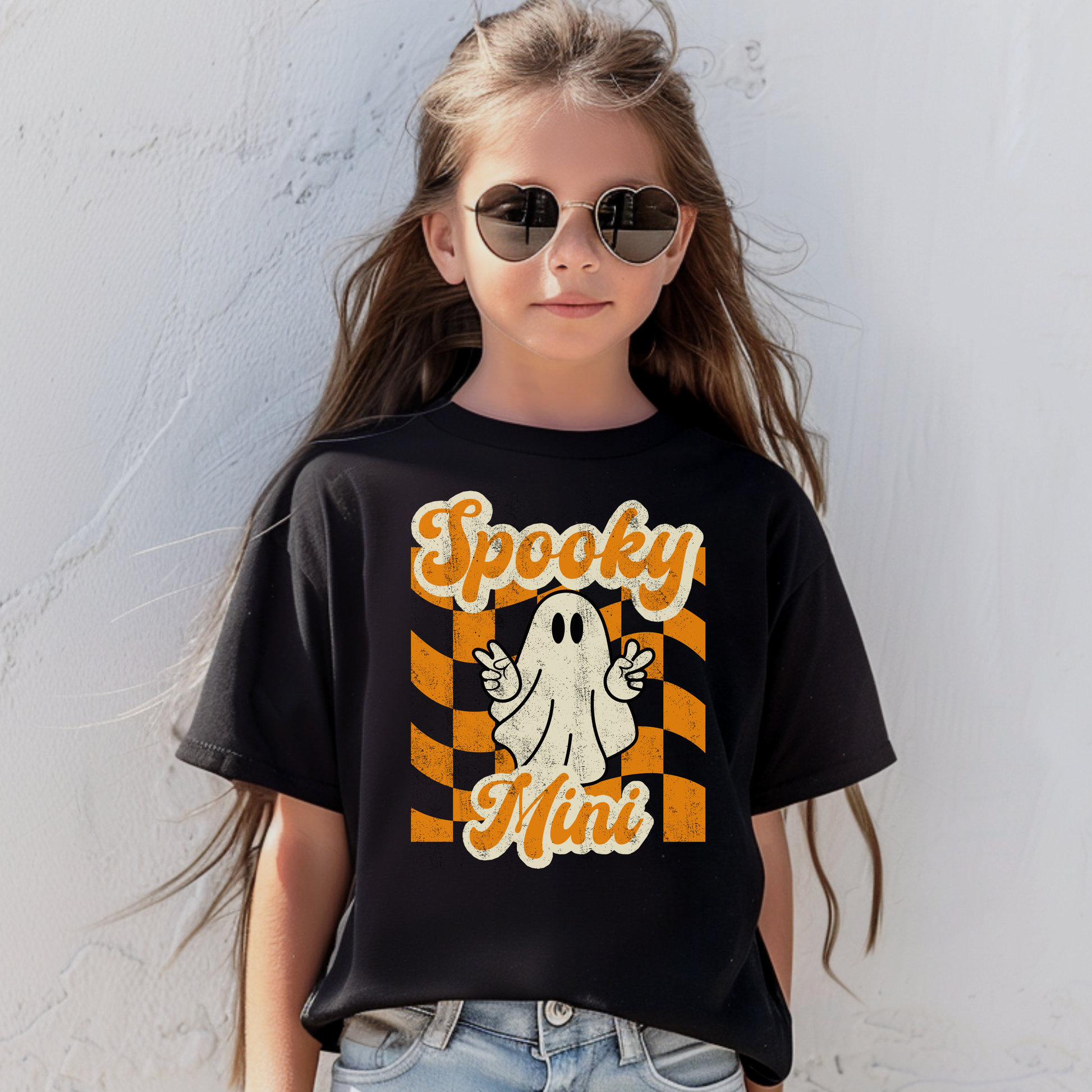 Black Bella Canvas 3001Y youth Halloween t shirt featuring Spooky Mini graphic with playful ghost making peace sign over orange checkered aesthetic background. Ideal for youth fall outfits, Halloween parties, school events, pumpkin patch trips, trick or treat night, and seasonal family matching outfits. Matches toddler Spooky Mini shirt and Spooky Mama adult tee for coordinated mommy and me Halloween clothing and family photo outfits.