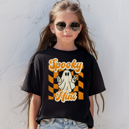 Black Bella Canvas 3001Y youth Halloween t shirt featuring Spooky Mini graphic with playful ghost making peace sign over orange checkered aesthetic background. Ideal for youth fall outfits, Halloween parties, school events, pumpkin patch trips, trick or treat night, and seasonal family matching outfits. Matches toddler Spooky Mini shirt and Spooky Mama adult tee for coordinated mommy and me Halloween clothing and family photo outfits.