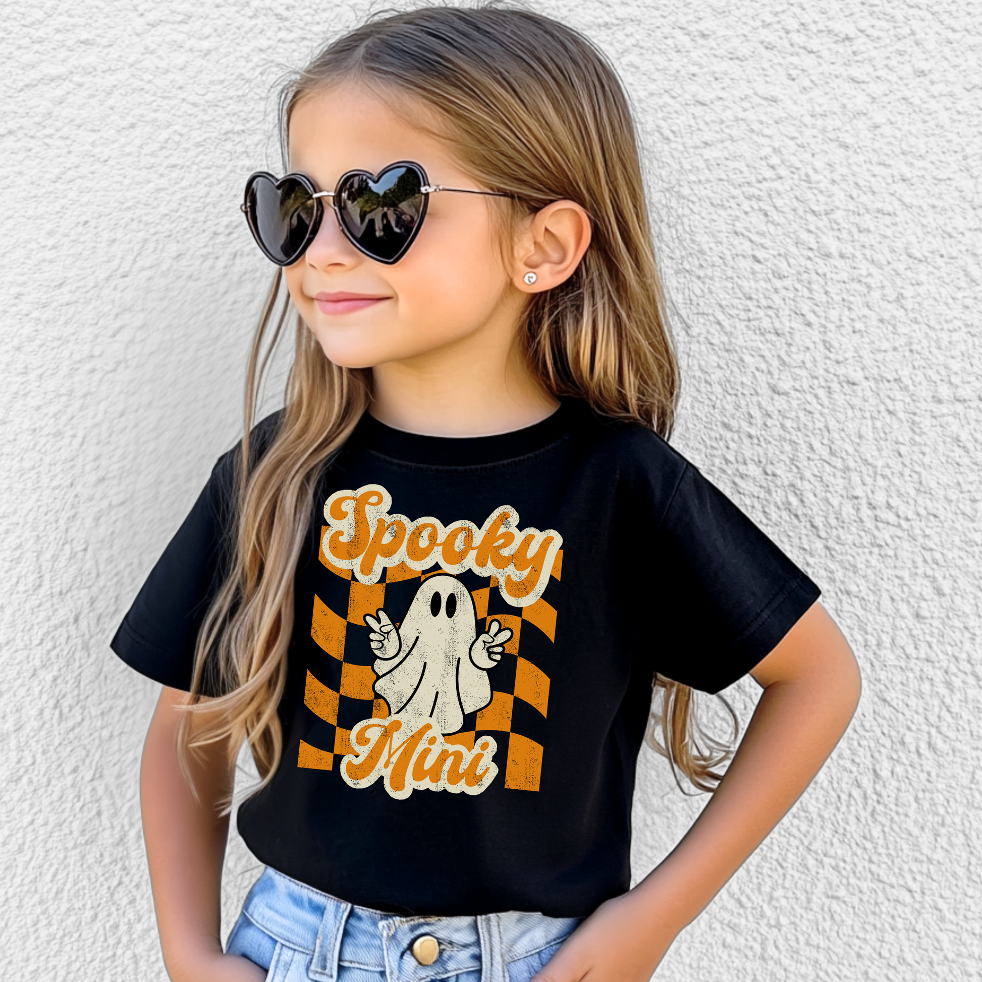 Spooky Mini youth Halloween shirt on black Bella Canvas 3001Y featuring adorable ghost with peace sign over orange checkered retro Halloween background. Perfect for youth spooky season outfits, school Halloween celebrations, fall festivals, pumpkin patch trips, and trick or treat night. Coordinates with toddler and adult matching shirts including Spooky Mini toddler tee and Spooky Mama adult Halloween shirt for family matching outfits.