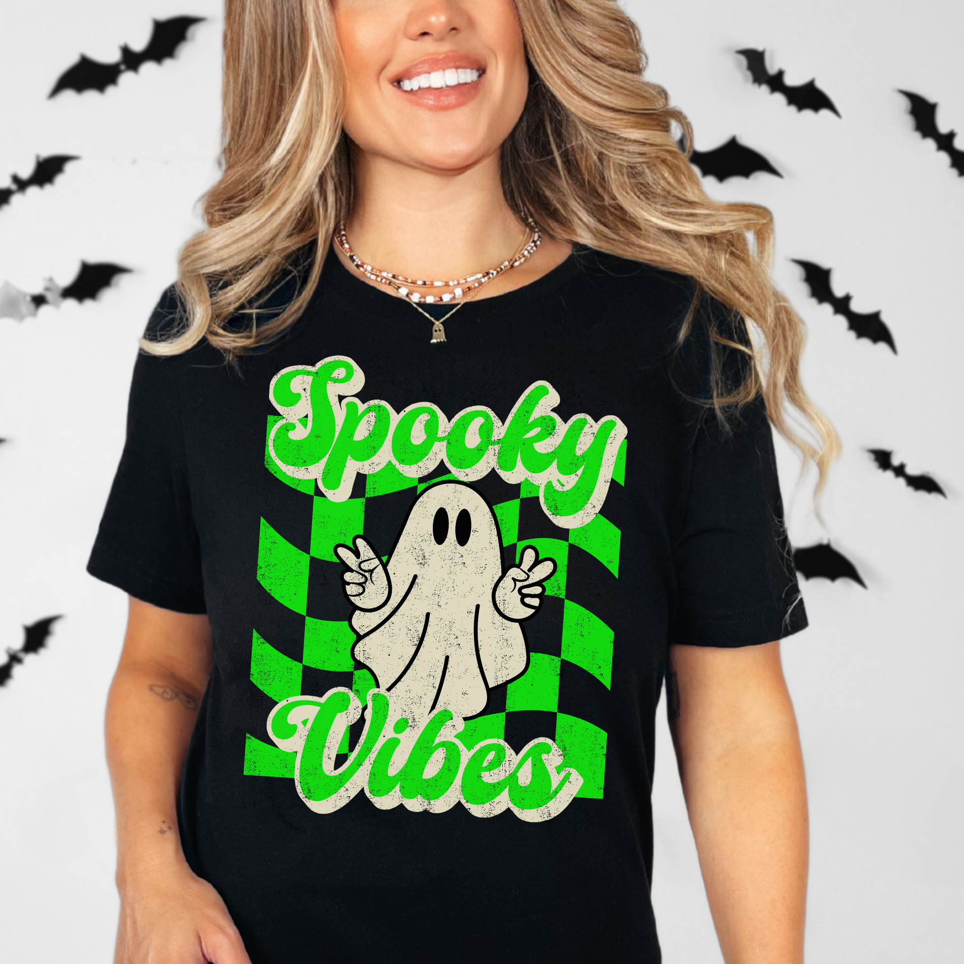 Black Bella Canvas 3001 Spooky Vibes Halloween shirt featuring a cute ghost giving peace signs over a bright green checkerboard background with retro spooky vibes lettering. Soft unisex graphic tee in sizes S to 3XL with matching child sizes for family matching Halloween outfits. Great for pumpkin patch trips, trick or treat night, classroom parties, haunted house visits, ghost lovers, spooky season style, October photos, and cute fall fashion looks with jeans or leggings.