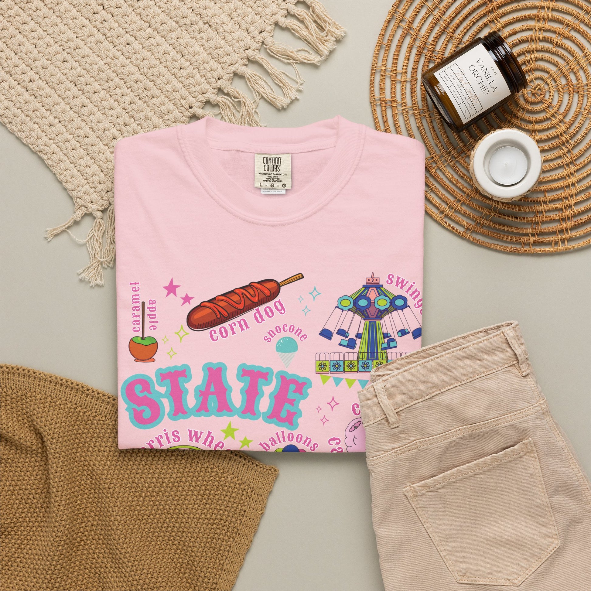 Flatlay of a colorful state fair T shirt graphic featuring the words STATE FAIR surrounded by classic midway imagery including a Ferris wheel, roller coaster, carnival ride, cotton candy, balloons, funnel cake, corn dogs, livestock, blue ribbon, tickets and sparkling stars at a county fair.