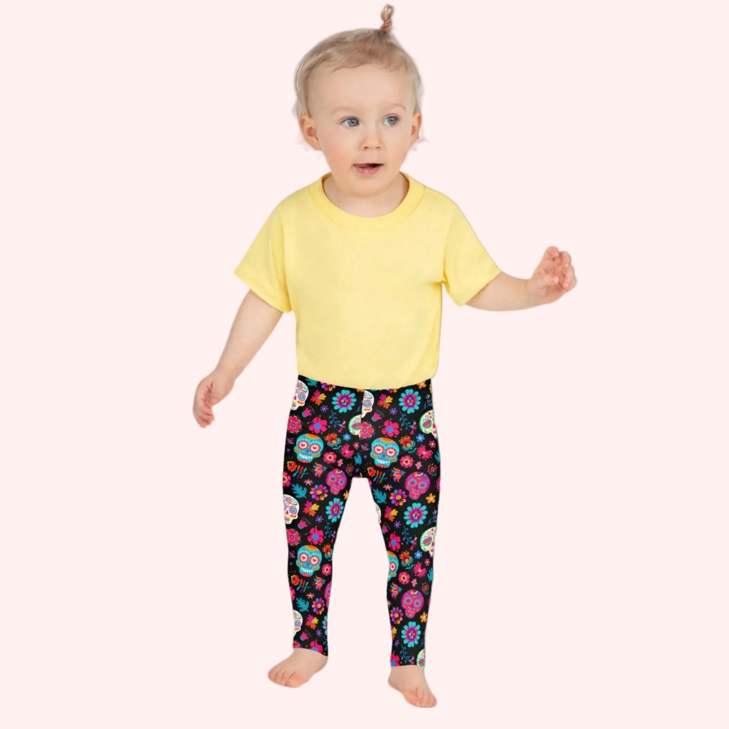 Toddler girl wearing colorful Dia de los Muertos leggings featuring vibrant sugar skulls, marigolds and colorful floral details against a bold black background. These girls Day of the Dead leggings have a bright all over print with pink, orange, yellow, blue, green and purple accents and a soft stretchy fit designed for comfortable movement during play, family celebrations, cultural events, fall festivals and everyday adventures.