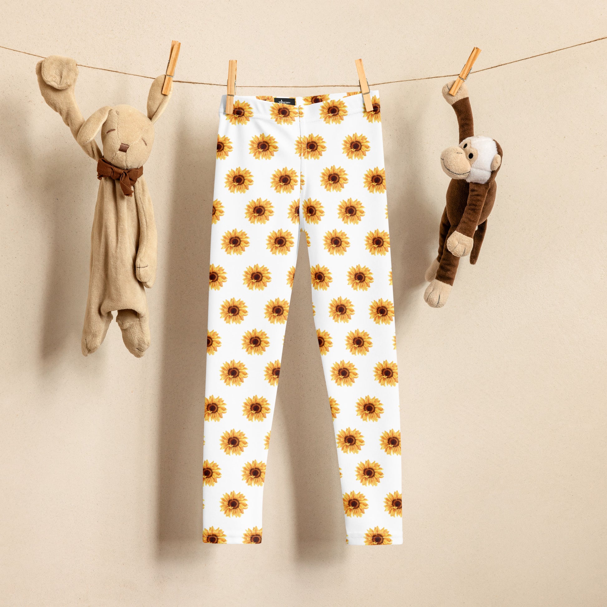 Adorable sunflower print toddler leggings with a bright yellow floral design on soft white fabric, shown hanging between plush toys. These high-rise, full-length leggings are perfect for active girls who love comfort and color. Ideal for school, playdates, or cozy days at home, they’re made from smooth, stretchy microfiber that won’t fade. A sweet sunflower outfit idea and part of a matching mommy-and-me and sister leggings collection.