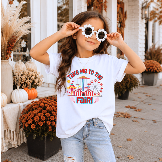 A young girl wearing a white graphic t-shirt that says 'TAKEN ME TO THE FAIR!' with a carnival fair illustration, paired with light-wash jeans. She is holding a pair of white flower-shaped sunglasses in front of her eyes, posing outdoors on a porch decorated for autumn.
