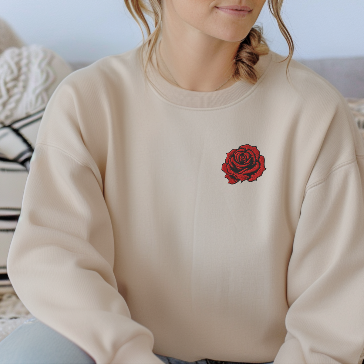 Woman wearing a sand colored crewneck sweatshirt with a red embroidered rose on the chest, styled in a bright neutral bedroom. The full length view shows the classic silhouette, long sleeves, and contrast between the warm neutral fabric and red floral embroidery. Women's embroidered rose sweatshirt with a timeless look for casual outfits, June birthdays, and thoughtful gifts.