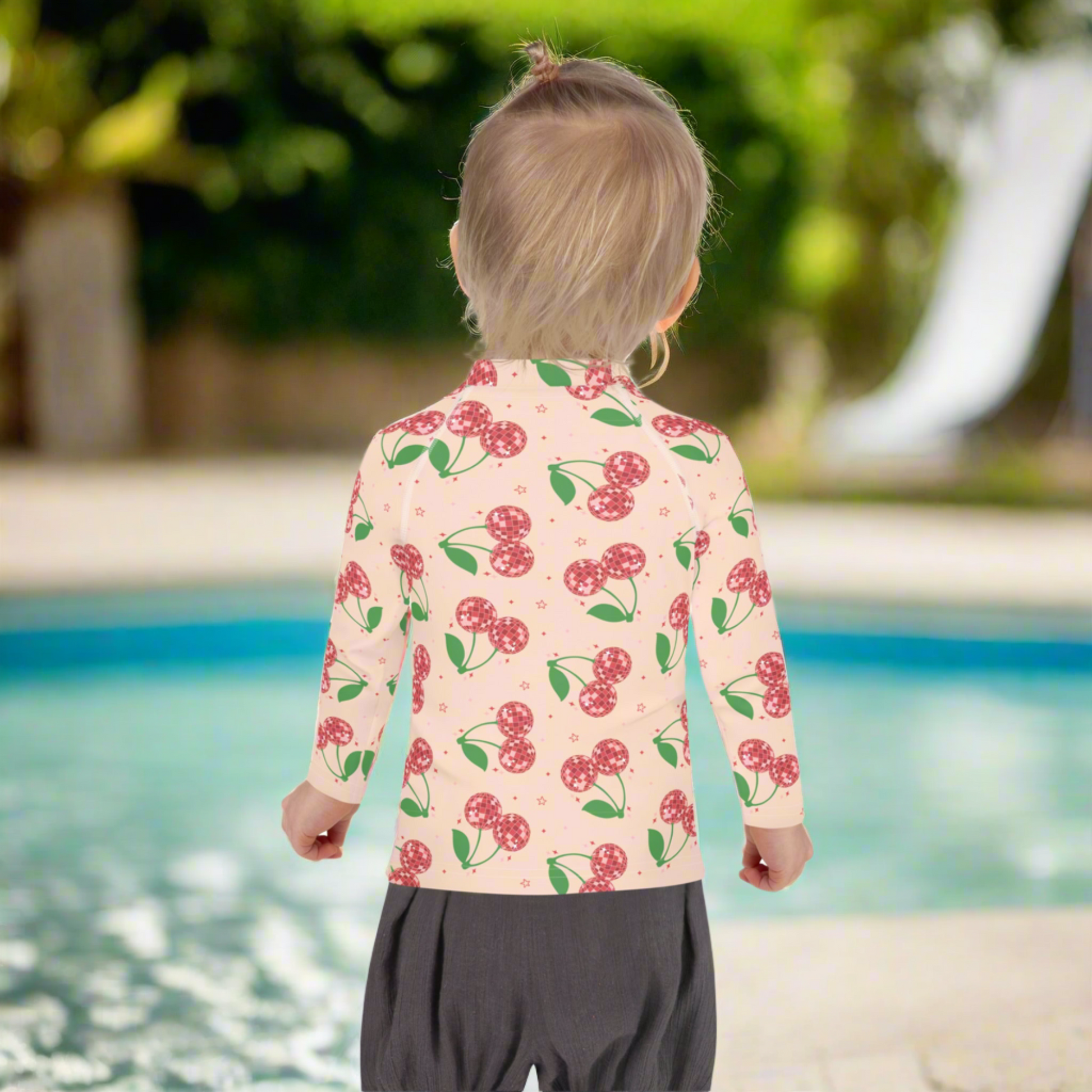 Toddler girls rash guard in a disco cherry print with a light pink background and bright red cherries, available in sizes 2T to 7. This long sleeve swim top features UPF 50 sun protection and flexible stretch fabric for comfort and movement. Perfect for beachwear, pool outfits, splash pad play, and summer travel. A fun and stylish option for kids swimwear, with matching mommy and me pieces available for coordinated family outfits.
