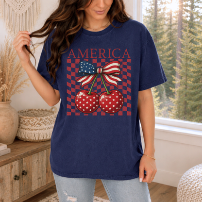 Woman wearing Navy Comfort Colors America Cherry Bow shirt styled for a casual patriotic summer look. Deep navy garment dyed tee features vintage America lettering above two glossy red cherries with white star details tied together by a bold stars and stripes bow. Distressed checkerboard background adds retro Americana charm and contrast. Relaxed unisex fit pairs perfectly with denim shorts, white jeans, or sneakers. Cute women’s patriotic shirt for July fourth, fireworks, parades, lake trips, concerts, and