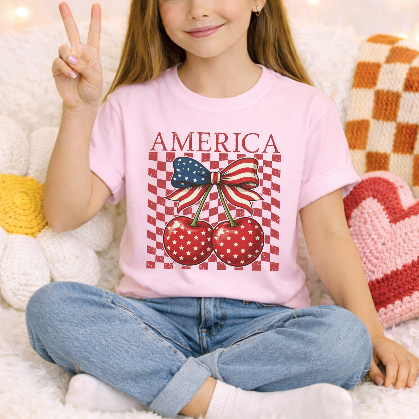 Blossom pink youth America Cherry Bow shirt designed on a Comfort Colors heavyweight tee. Front graphic features patriotic cherries with white stars, green stems, and a red white and blue ribbon bow. Distressed checkerboard detail adds retro boutique style. Soft cotton shirt for girls that pairs easily with denim shorts or sneakers. Perfect for patriotic holidays, summer photos, family trips, and matching sibling or mommy outfits.