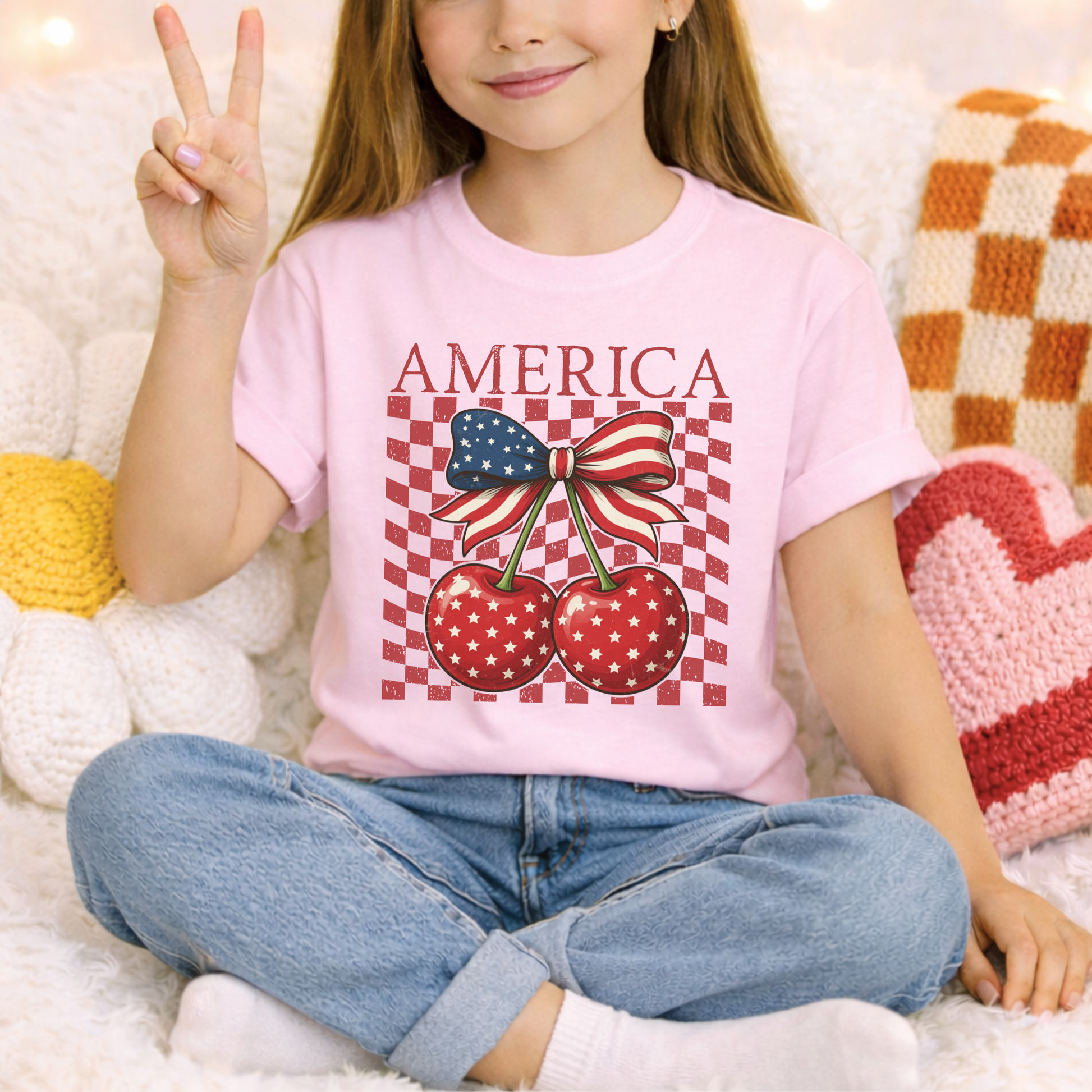 Blossom pink youth America Cherry Bow shirt designed on a Comfort Colors heavyweight tee. Front graphic features patriotic cherries with white stars, green stems, and a red white and blue ribbon bow. Distressed checkerboard detail adds retro boutique style. Soft cotton shirt for girls that pairs easily with denim shorts or sneakers. Perfect for patriotic holidays, summer photos, family trips, and matching sibling or mommy outfits.